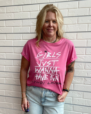A woman wearing a pink graphic t-shirt with white text that reads 'GIRLS JUST WANNA HAVE FUN -CYNDI LARBER' and light-wash distressed jeans, standing against a white brick wall.