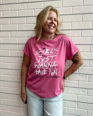 A woman wearing a pink graphic t-shirt with white text that reads 'GIRLS JUST WANNA HAVE FUN' and 'CLYNDI LAVER' underneath, paired with light-wash distressed jeans, standing against a white brick wall.