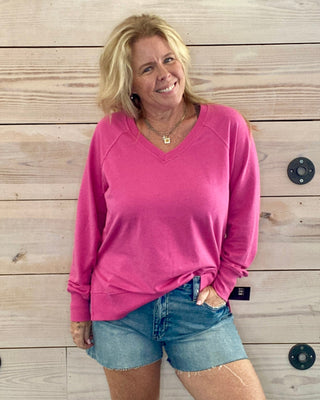 Woman wearing a pink sweater and denim shorts standing against a wooden wall.