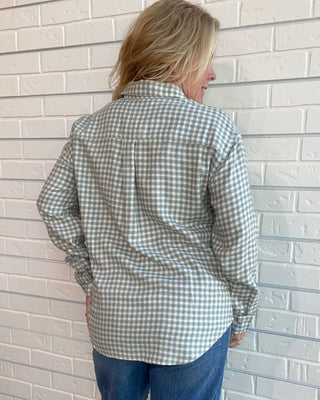 A woman viewed from behind wearing a light sage green and white gingham long-sleeve button-up shirt with blue jeans, standing against a white brick wall.