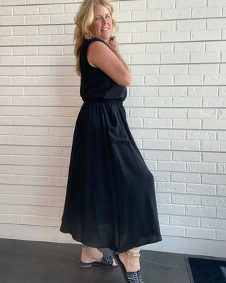 A woman modeling a sleeveless black midi skirt top set with a gathered waist, standing against a white brick wall.