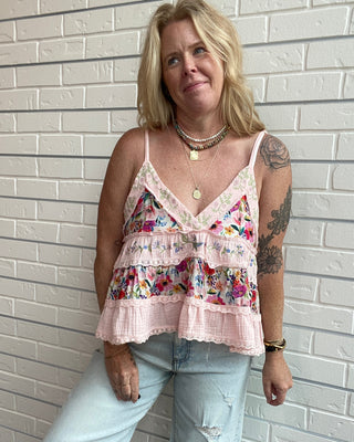 A woman with blonde hair and tattoos models a light pink floral and lace camisole top with light-wash distressed jeans, standing against a white brick wall.