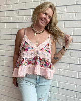 A woman wearing a light pink ruffled camisole top with a colorful floral print and light blue jeans, posing against a white brick wall.