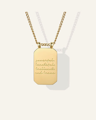 A gold rectangular pendant necklace with a chain, featuring the engraved words 'peaceful, beautiful, brilliant, and free' in a cursive font, displayed against a light cream background.