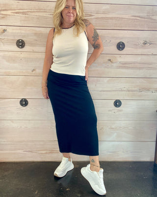 Woman wearing a white tank top and black skirt standing against a wooden wall.