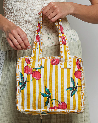 A person holds a small fabric tote bag with yellow and white vertical stripes and a cherry print. The person is wearing a textured cream top and a light olive green skirt against a plain light gray background.