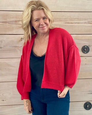 Woman wearing a red cardigan over a black top and blue jeans, standing against a wooden wall.