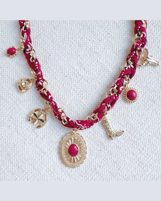 A western-themed charm necklace featuring a magenta and white patterned braided chain, gold-tone chain links, and various dangling charms including a cowboy boot, an oval medallion with a pink stone, and a small pink gem, displayed on a textured white fabric background.