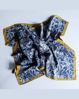 A square scarf with a navy blue and white paisley block print pattern, finished with a contrasting gold satin border, laid out on a light gray background.
