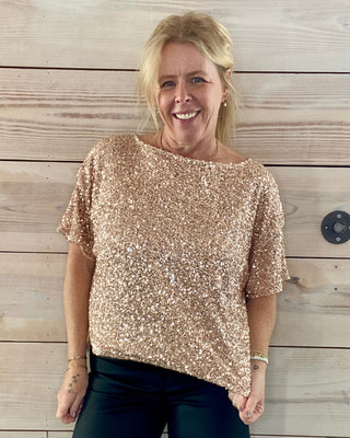 Woman wearing a gold sequin top and black pants standing against a wooden wall.