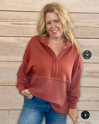 Woman wearing a rust-colored pullover and blue jeans standing against a wooden wall.
