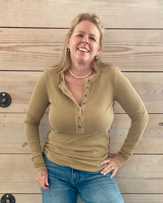 Woman wearing a green long-sleeve shirt and blue jeans standing against a wooden wall.