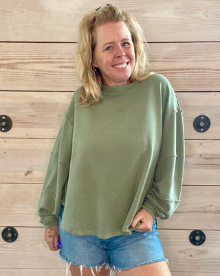 Woman wearing a green sweater and denim shorts standing against a wooden wall.