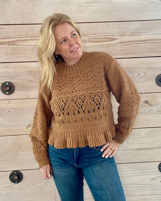 Woman wearing a brown knitted sweater and blue jeans standing on a wooden floor.