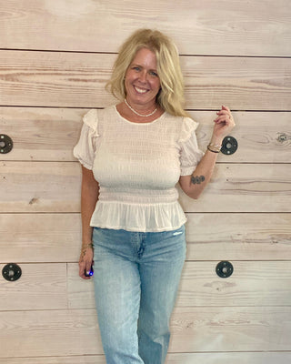 Woman wearing a white blouse and blue jeans standing against a wooden wall.
