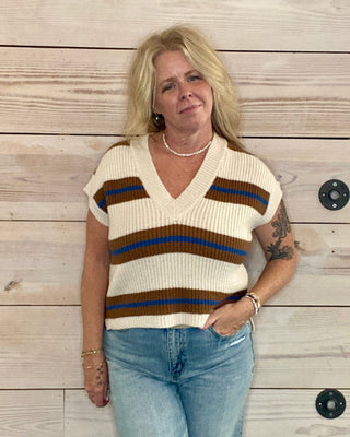 Woman wearing a striped sweater vest and jeans against a wooden wall.