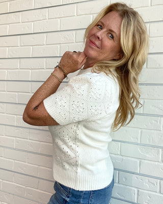 A blonde woman wearing a white short-sleeve knit sweater and blue jeans, posing against a white brick wall.