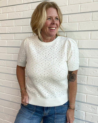 A smiling blonde woman wearing a short-sleeve cream knit sweater with a textured eyelet pattern and blue jeans, standing against a white painted brick wall.