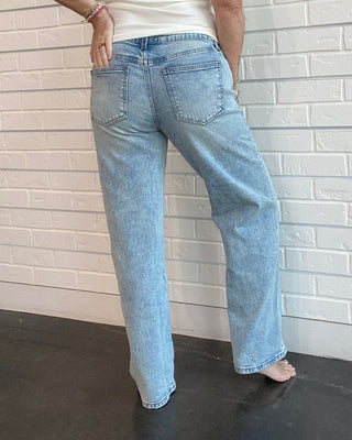 Person wearing a white sleeveless top and light blue jeans against a white brick wall.