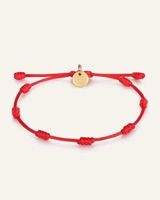 A red adjustable cord bracelet with knotted beads and a small round gold disc charm, displayed on a light cream background.