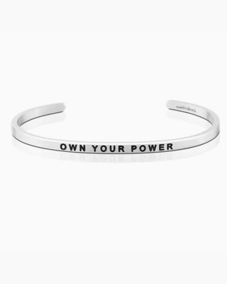 A silver-toned metal cuff bracelet with the words 'OWN YOUR POWER' engraved in black capital letters on the front, shown on a clean white background.
