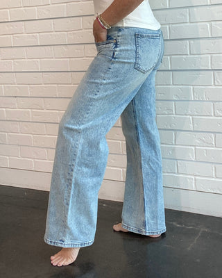 Woman wearing a white tank top and light blue jeans standing against a white brick wall.