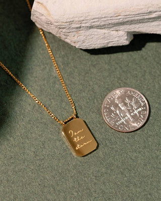 Gold necklace with a dog tag and engraved text, next to a U.S. quarter on a green surface.