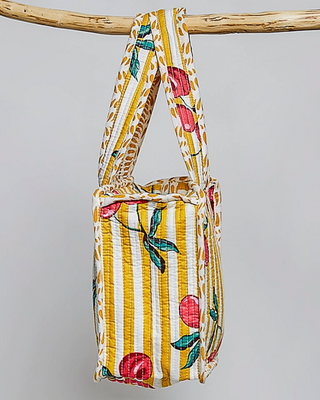A small fabric tote bag with yellow and white vertical stripes and a red fruit and green leaf print, hanging from a natural wood branch against a light gray background.