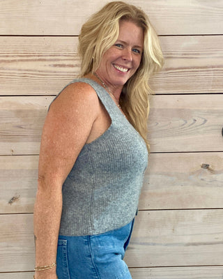Woman wearing a sleeveless top and jeans standing against a wooden wall.