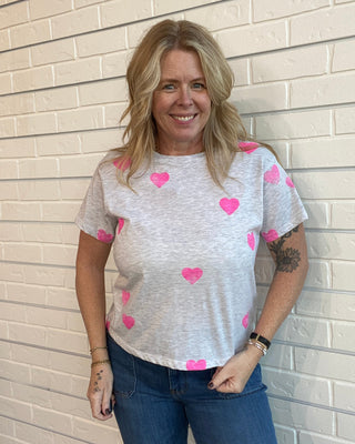 Woman wearing a white top with pink hearts and blue jeans against a tiled wall.