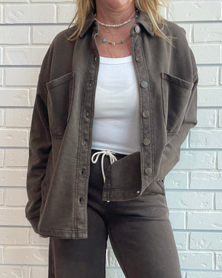 A woman wearing a brown washed button-up shirt jacket over a white tank top, paired with matching brown wide-leg jeans and leopard print shoes, standing against a white brick wall.