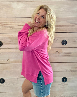 Woman wearing a pink long-sleeve top and denim shorts against a wooden wall.