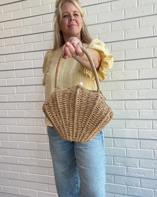 Woman holding a woven handbag against a brick wall.