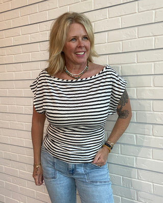 Woman wearing a black and white striped shirt and blue jeans standing against a light brick wall.
