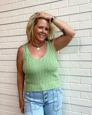 Woman wearing a green sleeveless top and blue jeans against a white brick wall.