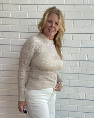 A smiling blonde woman wearing a beige textured knit mock-neck sweater and white pants, standing against a white brick wall.