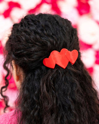 Person with wavy hair wearing a red heart-shaped hair clip against a blurred pink and white background
