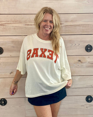 Woman wearing a white sweatshirt with 'TEXAS' printed on it against a wooden wall.