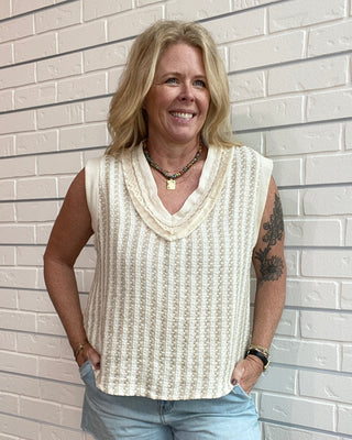 Woman wearing a sleeveless beige top and denim shorts standing against a light brick wall.