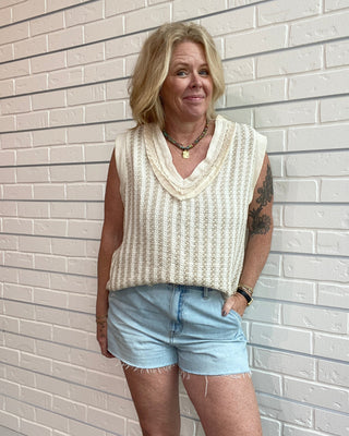 Woman wearing a sleeveless beige top and light blue shorts standing against a white brick wall.
