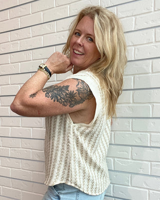 Woman wearing a sleeveless patterned top and denim shorts against a white brick wall.