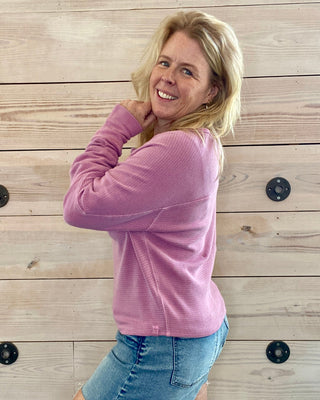 Woman wearing a pink sweater and denim skirt against a wooden wall.