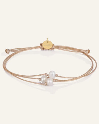A delicate multi-strand cord bracelet featuring three white freshwater pearls and a small round gold charm engraved with 'mantra', displayed on a clean off-white background.