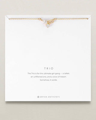 A gold-tone tri-tera necklace with a clear crystal charm, presented on a white backing card with the name 'TRIO' and a quote by Bryan Anthony's.