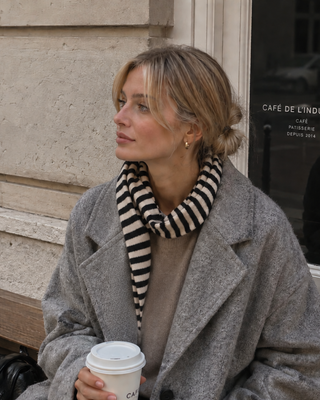 Woman in a gray coat with a striped scarf holding a coffee cup, sitting on steps in front of a building.