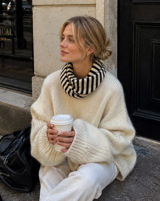 Woman in a cream sweater and striped scarf holding a coffee cup, sitting on steps.
