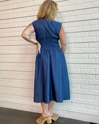 Woman wearing a blue dress standing facing t a white brick wall.