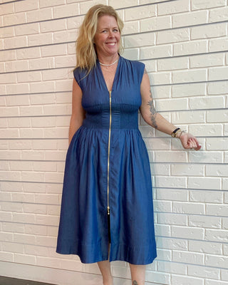 Woman wearing a blue dress with a zipper standing against a white brick wall.