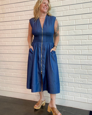 Woman wearing a blue dress standing against a light brick wall.