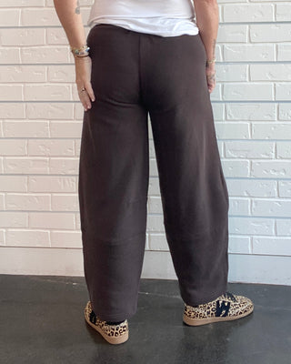 A woman viewed from behind wearing loose dark brown wide-leg pants, a white sleeveless top, and leopard-print shoes, standing against a white brick wall.